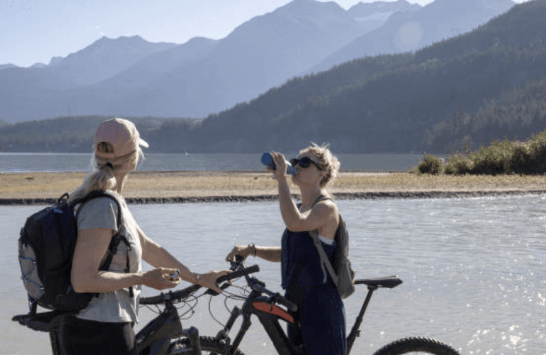 Two women with electric bikes resting near a lake, mountains in background, one drinking water