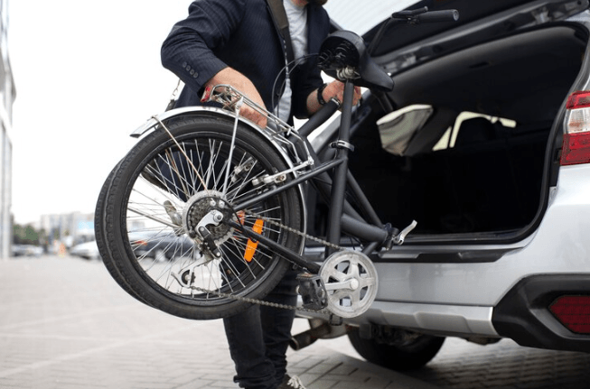 Folded black electric bike being loaded into a car trunk, side view, urban setting