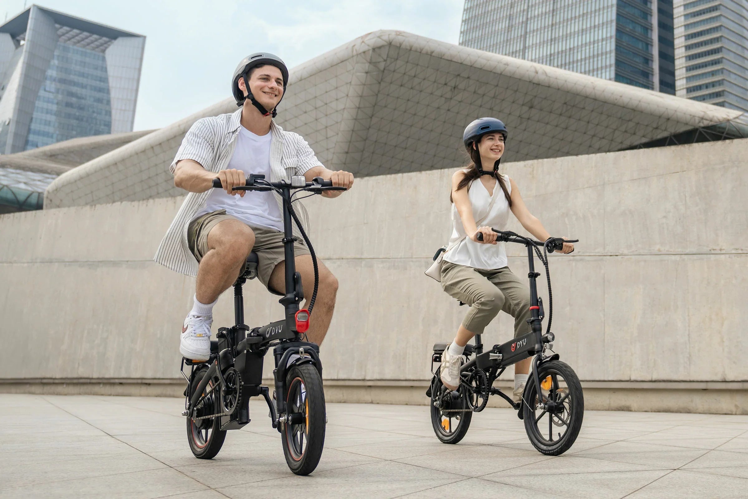 Two people riding DYU A5 Upgraded Folding Electric Bikes in an urban setting with modern buildings in the background.